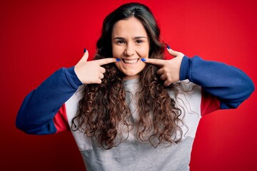 Sticker - Young beautiful woman with curly hair wearing casual sweatshirt over isolated red background Smiling with open mouth, fingers pointing and forcing cheerful smile