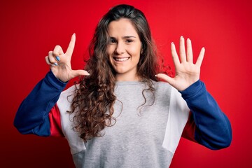 Sticker - Young beautiful woman with curly hair wearing casual sweatshirt over isolated red background showing and pointing up with fingers number seven while smiling confident and happy.