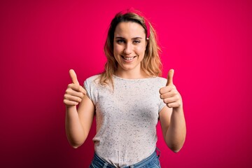 Young beautiful blonde woman wearing casual t-shirt standing over isolated pink background success sign doing positive gesture with hand, thumbs up smiling and happy. Cheerful expression.