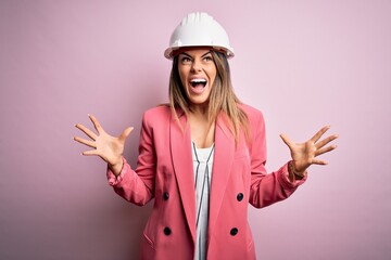 Canvas Print - Young beautiful brunette architect woman wearing safety helmet over pink background crazy and mad shouting and yelling with aggressive expression and arms raised. Frustration concept.