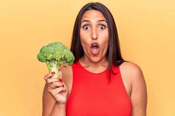 Sticker - Young beautiful brunette woman holding broccoli scared and amazed with open mouth for surprise, disbelief face