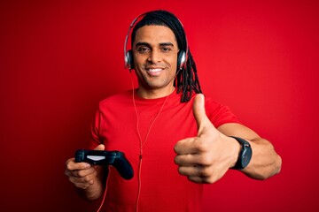 Wall Mural - African american man with dreadlocks playing video game using joystick and headphones happy with big smile doing ok sign, thumb up with fingers, excellent sign