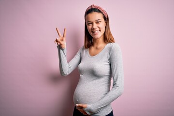 Poster - Young beautiful teenager girl pregnant expecting baby over isolated pink background smiling with happy face winking at the camera doing victory sign with fingers. Number two.