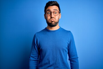 Canvas Print - Young handsome man with beard wearing casual sweater and glasses over blue background puffing cheeks with funny face. Mouth inflated with air, crazy expression.