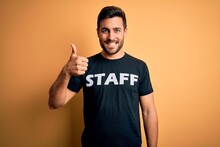Young Handsome Worker Man With Beard Wearing Staff Uniform T-shirt Over Yellow Background Happy With Big Smile Doing Ok Sign, Thumb Up With Fingers, Excellent Sign