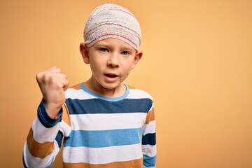 Wall Mural - Young little caucasian kid injured wearing medical bandage on head over yellow background angry and mad raising fist frustrated and furious while shouting with anger. Rage and aggressive concept.