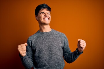 Poster - Young handsome man wearing casual sweater standing over isolated orange background very happy and excited doing winner gesture with arms raised, smiling and screaming for success. Celebration concept.