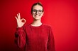 Young beautiful redhead woman wearing casual sweater over isolated red background smiling positive doing ok sign with hand and fingers. Successful expression.