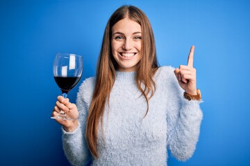 Young beautiful redhead woman drinking glass of red wine over isolated blue background surprised with an idea or question pointing finger with happy face, number one