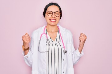 Wall Mural - Beautiful doctor woman with blue eyes wearing coat and stethoscope over pink background excited for success with arms raised and eyes closed celebrating victory smiling. Winner concept.