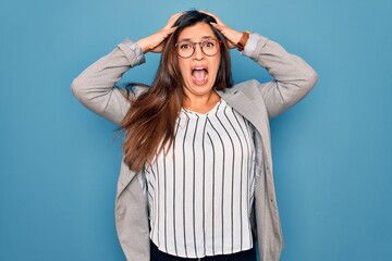 Sticker - Young hispanic business woman wearing glasses standing over blue isolated background Crazy and scared with hands on head, afraid and surprised of shock with open mouth