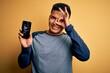 © Krakenimages.com - Young handsome latin man holding broken smartphone showing craked screen with happy face smiling doing ok sign with hand on eye looking through fingers