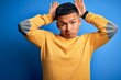© Krakenimages.com - Young handsome latin man wearing yellow casual sweater over isolated blue background Doing bunny ears gesture with hands palms looking cynical and skeptical. Easter rabbit concept.