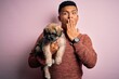 © Krakenimages.com - Young handsome latin man holding cute puppy pet over isolated pink background cover mouth with hand shocked with shame for mistake, expression of fear, scared in silence, secret concept