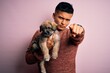 © Krakenimages.com - Young handsome latin man holding cute puppy pet over isolated pink background pointing with finger to the camera and to you, hand sign, positive and confident gesture from the front