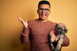© Krakenimages.com - Young handsome latin man holding cute puppy pet over isolated yellow background very happy and excited, winner expression celebrating victory screaming with big smile and raised hands