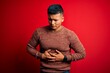 © Krakenimages.com - Young handsome latin man wearing casual sweater standing over red background with hand on stomach because indigestion, painful illness feeling unwell. Ache concept.