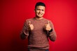 © Krakenimages.com - Young handsome latin man wearing casual sweater standing over red background success sign doing positive gesture with hand, thumbs up smiling and happy. Cheerful expression and winner gesture.