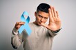 © Krakenimages.com - Young handsome latin man holding blue cancer ribbon over isolated white background with open hand doing stop sign with serious and confident expression, defense gesture
