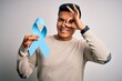 © Krakenimages.com - Young handsome latin man holding blue cancer ribbon over isolated white background with happy face smiling doing ok sign with hand on eye looking through fingers