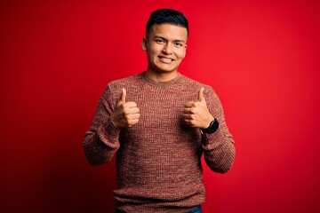 Young handsome latin man wearing casual sweater standing over red background success sign doing positive gesture with hand, thumbs up smiling and happy. Cheerful expression and winner gesture.