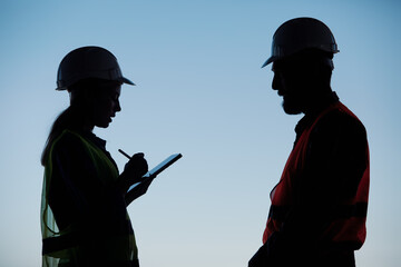 Wall Mural - Silhouettes of an engineer with a tablet in his hands and a construction worker in a hard hat against the blue sky stand opposite each other