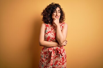 Poster - Young beautiful curly arab woman on vacation wearing summer floral dress and sunglasses with hand on chin thinking about question, pensive expression. Smiling with thoughtful face. Doubt concept.