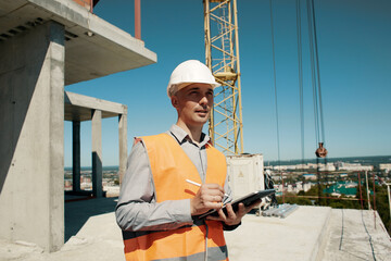 An engineer in an orange vest and a white construction control helmet conducts an inspection with a tablet in his hands against the background of a construction site and a tower crane