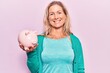 © Krakenimages.com - Middle age caucasian blonde woman holding piggy bank looking positive and happy standing and smiling with a confident smile showing teeth