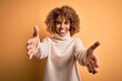 Young beautiful african american woman wearing turtleneck sweater over yellow background looking at the camera smiling with open arms for hug. Cheerful expression embracing happiness.