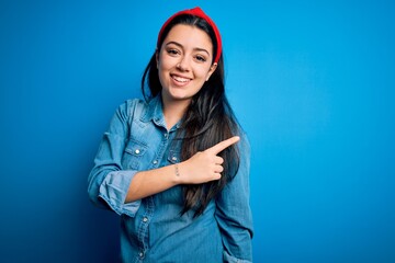 Sticker - Young brunette woman wearing casual denim shirt over blue isolated background cheerful with a smile on face pointing with hand and finger up to the side with happy and natural expression