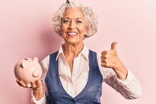 Senior Grey-haired Woman Holding Piggy Bank Smiling Happy And Positive, Thumb Up Doing Excellent And Approval Sign
