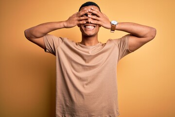 Wall Mural - Young handsome african american man wearing casual t-shirt standing over yellow background covering eyes with hands smiling cheerful and funny. Blind concept.