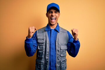 Wall Mural - Young handsome african american handyman wearing worker uniform and cap celebrating surprised and amazed for success with arms raised and open eyes. Winner concept.