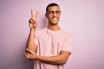 Wall Mural - Handsome african american man wearing casual t-shirt and glasses over pink background smiling with happy face winking at the camera doing victory sign with fingers. Number two.