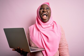 Poster - African american plus size woman wearing muslim hijab using laptop over pink background angry and mad screaming frustrated and furious, shouting with anger. Rage and aggressive concept.