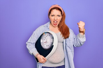 Wall Mural - Young beautiful redhead pregnant woman expecting baby holding weighting machine annoyed and frustrated shouting with anger, crazy and yelling with raised hand, anger concept