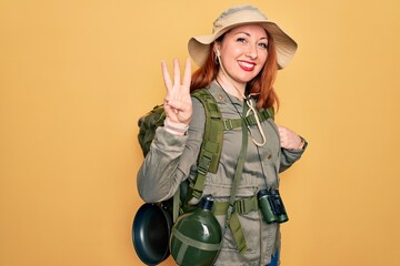 Poster - Young redhead backpacker woman hiking wearing backpack and hat over yellow background showing and pointing up with fingers number three while smiling confident and happy.