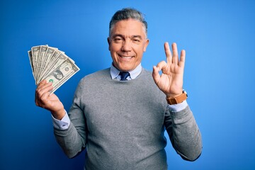 Sticker - Middle age handsome grey-haired man holding bunch of dollars over blue background doing ok sign with fingers, excellent symbol