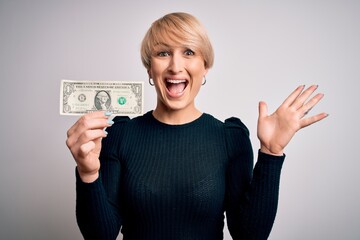 Wall Mural - Young blonde woman with short hair holding one dollar banknote over isolated background very happy and excited, winner expression celebrating victory screaming with big smile and raised hands