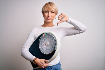 Sticker - Young blonde woman with short hair holding scale for healthy weight and lifestyle with angry face, negative sign showing dislike with thumbs down, rejection concept