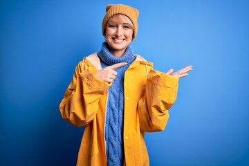 Sticker - Young blonde woman with short hair wearing rain coat for rainy weather over blue background amazed and smiling to the camera while presenting with hand and pointing with finger.