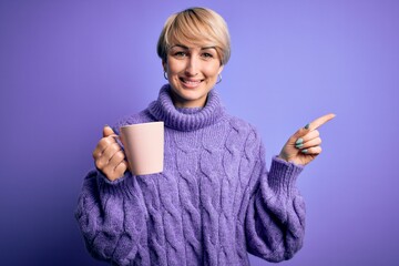 Poster - Young blonde woman with short hair wearing winter sweater drinking a cup of hot coffee very happy pointing with hand and finger to the side