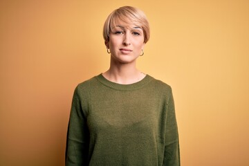 Poster - Young blonde woman with modern short hair wearing casual sweater over yellow background Relaxed with serious expression on face. Simple and natural looking at the camera.