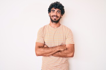 Handsome young man with curly hair and bear wearing casual striped tshirt happy face smiling with crossed arms looking at the camera. positive person.