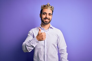 Sticker - Young handsome man with beard wearing golden crown of king over purple background doing happy thumbs up gesture with hand. Approving expression looking at the camera showing success.