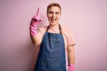 Wall Mural - Young handsome redhead cleaner man doing housework wearing apron and gloves pointing finger up with successful idea. Exited and happy. Number one.