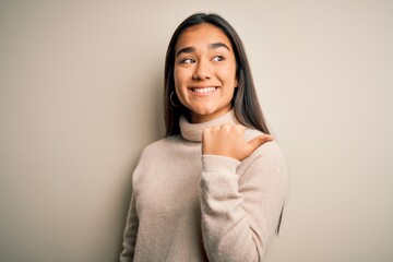 Canvas Print - Young beautiful asian woman wearing casual turtleneck sweater over white background smiling with happy face looking and pointing to the side with thumb up.