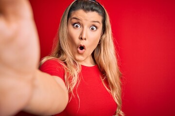 Sticker - Young beautiful blonde woman making selfie by the camer over isolated red background scared in shock with a surprise face, afraid and excited with fear expression