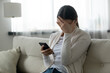 © fizkes - Woman sit on sofa holding smartphone cover face with hand feels scared humiliated suffering from cyberbullying being on-line abused by stalker. Bad news, life troubles, break up with boyfriend concept